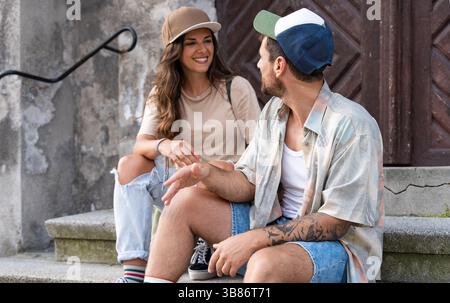 Young happy hipster millennial gen z couple in love and modern clothes sitting outdoor on the street talking and having fun. Man and woman university Stock Photo