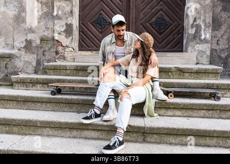 Young happy hipster millennial gen z couple in love and modern clothes sitting outdoor on the street talking and having fun. Man and woman university Stock Photo