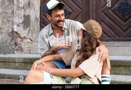 Young happy hipster millennial gen z couple in love and modern clothes sitting outdoor on the street talking and having fun. Man and woman university Stock Photo
