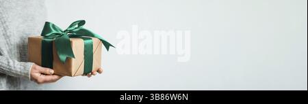 Female hands hold gift box on white background, copy space right. Unrecognizable caucasian girl in gray sweater holding gift box in craft wrapping pap Stock Photo