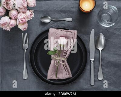 Beautiful table setting with candle on gray linen tablecloth. Festive table setting for wedding dinner with pink spray rose and pink napkin on plate. Stock Photo