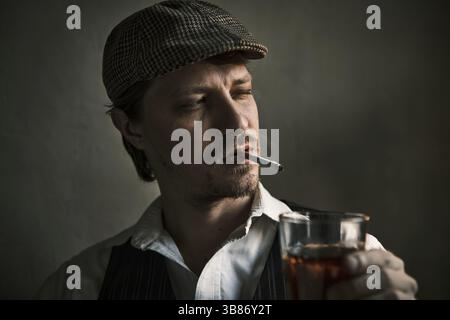 Young guy smokes a cigarette and drinks whiskey, blows smoke, dressed in a retro style, in a beret, cinematic shot, big closeup Stock Photo