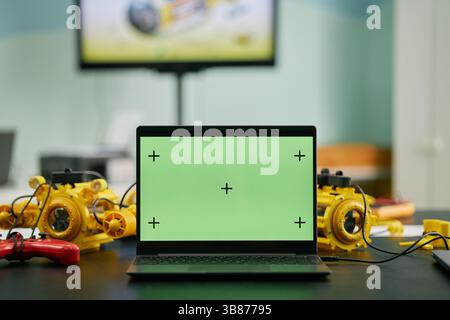 Scene displaying a laptop with a green screen on table surrounded by various robotic devices. Background includes blurry television screen showing rob Stock Photo