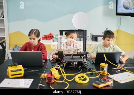 Students engaging with 3D printer and laptops, focused on project. Varied expressions show concentration. Classroom decorated with educational visuals Stock Photo