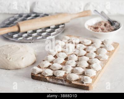 Uncooked russian pelmeni on cutting board and ingredients for homemade pelmeni on white table. Process of making pelmeni, ravioli or dumplings with me Stock Photo