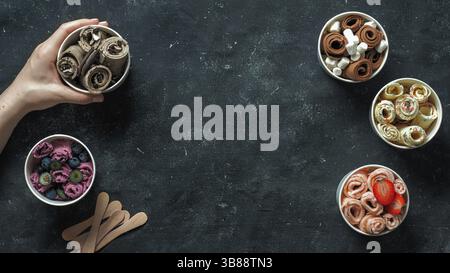 Hand with rolled ice cream in cone cup on dark background. Different iced rolls top view or flat lay. Thai style rolled ice cream with copy space in c Stock Photo