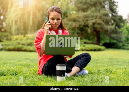 Young woman multitasking with laptop and smartphone while sitting on grass in city park Stock Photo