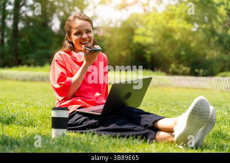 Young woman recording voice message while relaxing with laptop in city park. Digital communication. Stock Photo