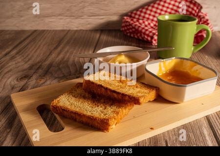 Toasted bread slices served on wooden cutting board with butter, apricot jam and coffee mug, ready for breakfast Stock Photo
