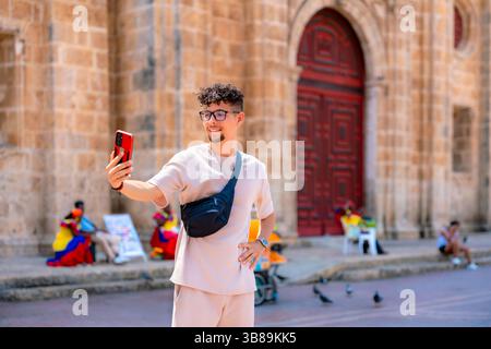 Young Latin man in beige streetwear smiling while using his phone near historic church in Cartagena, Colombia on a sunny day. Stock Photo