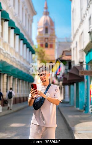 Confident young Latino man in beige outfit smiles while using phone on a vibrant colonial street in Cartagena, Colombia. Stock Photo