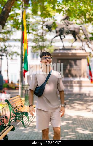 Stylish young Latino man in beige outfit and glasses walks confidently through a colorful sunny plaza in Cartagena, Colombia. Stock Photo