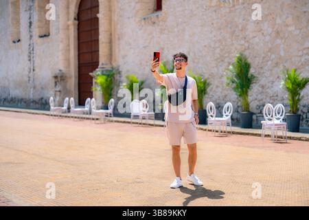 Latino man wearing beige outfit and sunglasses takes a cheerful selfie outside a historic building in Cartagena, Colombia. Stock Photo
