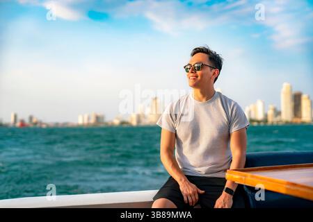 Smiling young Latino man sits on a boat near Cartagena, Colombia, wearing a gray shirt and black shorts with the city skyline behind him. Stock Photo
