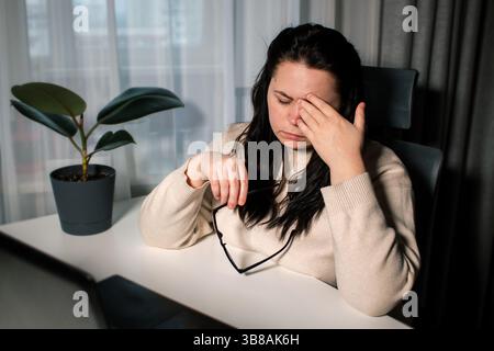 Symptoms of overworking. Young tired female freelancer in eyeglasses sitting in front of computer rubbing eyes feeling eye strain during work. Stock Photo