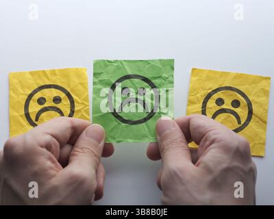 A man holding a green paper note with a sad face on it in his hands over other ones. Close up. Stock Photo
