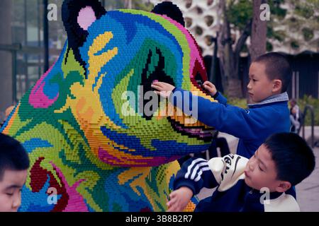 Children curiously touching a colorful, pixelated panda sculpture made from LEGO bricks Stock Photo