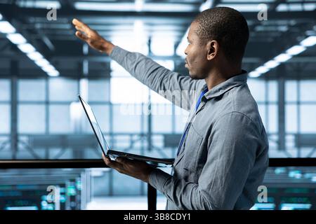 Technician holding laptop, looking around data center, managing server virtualization, finetuning network protocols, verifying high availability configurations, enforcing advanced security measures Stock Photo