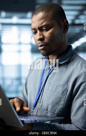 Computer scientist in data center using laptop to develop disaster recovery plans. African american man in server hub testing failover scenarios using notebook, protecting data during disruptions Stock Photo