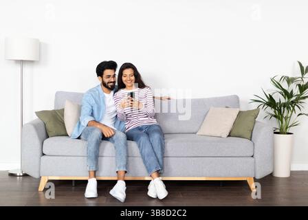Relaxed multicultural couple sitting on sofa, using smartphone Stock Photo