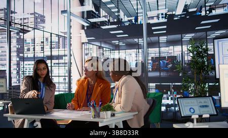 Diverse business experts sharing ideas for corporate growth planning, analyzing infographics for monthly report task. Powerful women working together to ensure organizational productivity. Camera B. Stock Photo