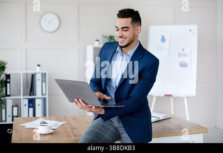 Cheerful young Arab businessman in formal suit using laptop computer, working online at modern office Stock Photo
