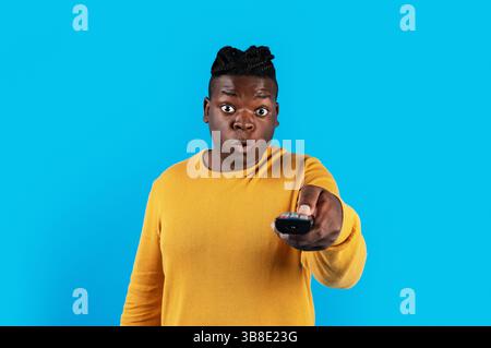 Shocked Black Man Watching Television, Pointing Remote Controller At Camera Stock Photo