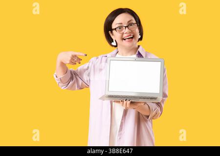Mature woman pointing at blank laptop on yellow background Stock Photo