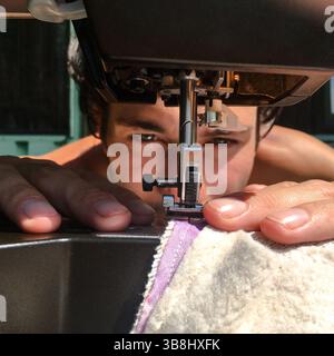 A close-up image of sewing machine's presser foot in foreground, while handsome young man  sewing fabrics together. Hobby sewing by full concentration Stock Photo
