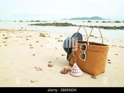 Summer woven bag with blue straw hat and sunglasses on the sandy beach with natural seashells Stock Photo