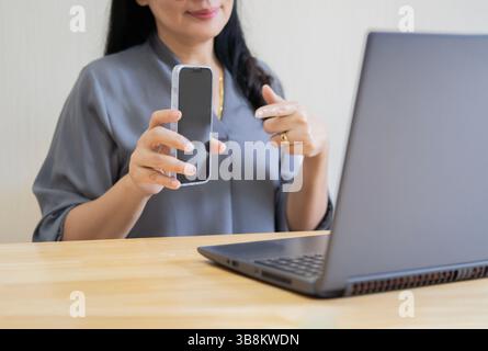 Attractive happy adult Asian woman sitting at the desk, using laptop computer, holding mobile phone. Selective focus. Stock Photo