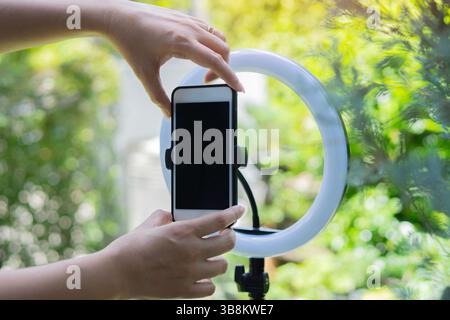 Selective focus of hands of influencer adjusting smart phone on ring light in the garden at home. Broadcast outdoor concept. Stock Photo