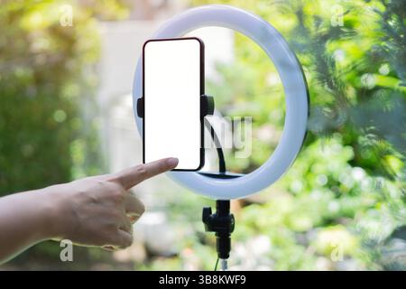 Woman's hand reach for the phone with blank screen, fixed on a round lamp with a tripod over nature background. Side view, selective focus. Stock Photo