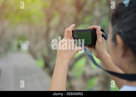 Back view of young cheerful woman traveler holds a camera in her hands while standing on the natural park. Copy space, selective focus. Stock Photo