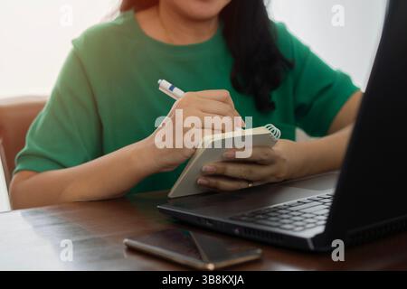 Cropped view and close up of woman's hands using laptop and writing in notepad. Working on project concept Stock Photo