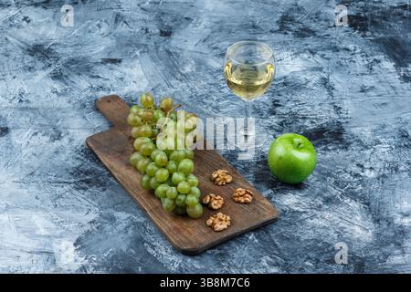 high angle view white grapes, walnuts on cutting board with glass of whisky, green apple on dark blue marble background. horizontal Stock Photo