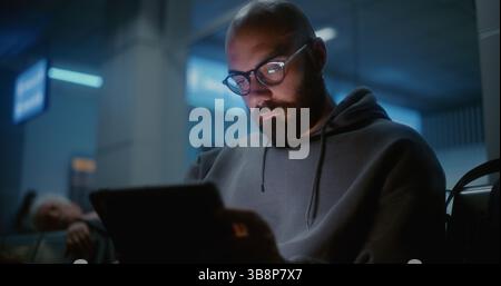 Crowded International Airport Terminal: Tired and Sleepy Man Yawning, Falling Asleep When Using Digital Tablet. Passenger Sitting in Boarding Lounge of Airline Hub at Night, Waiting for Delayed Flight Stock Photo