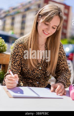 Blonde woman writing in notebook at outdoor cafe, sunlight warming surroundings, radiating cheerful urban lifestyle and relaxed atmosphere Stock Photo