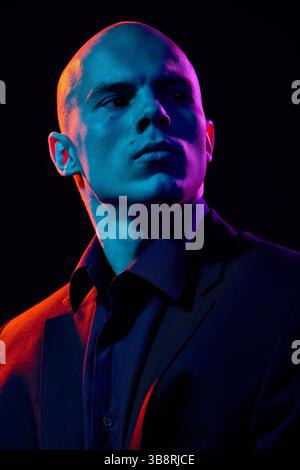 Close-up portrait of handsome bald man in dark shirt and jacket,posing confidently on dark background in multi-colored neon light Stock Photo