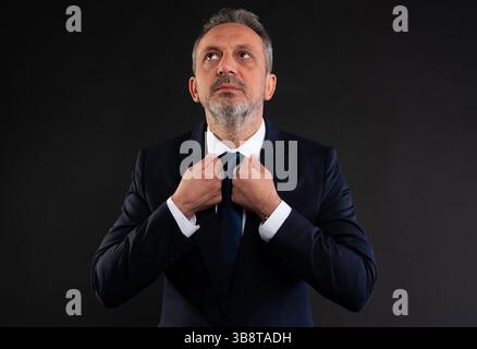 Middle-aged man in a suit adjusting his tie while looking upward, symbolizing ambition, hope, vision or reflection in a corporate setting. Stock Photo