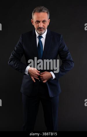 Mature businessman adjusting his formal outfit, buttoning his navy suit jacket while looking confidently at the camera. Stock Photo