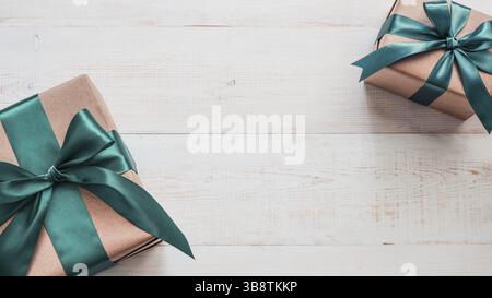 Two gift boxes in craft wrapping paper and green satin ribbon on white wooden table, copy space in center. Beautiful Christmas, New Year or Birthday p Stock Photo