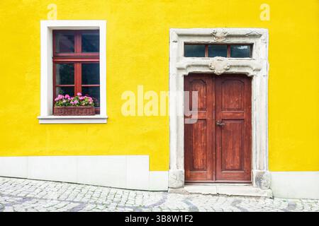 Old wooden door. Yellow house wall. Red window with flowers. Vintage entrance style. European architecture background. Bright colors historic charm. Stock Photo