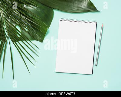 Blank paper notebook with pencil. Empty paper sketchbook with pincil and tropical leaves on blue background. Top view or flat lay. Copy space for text Stock Photo