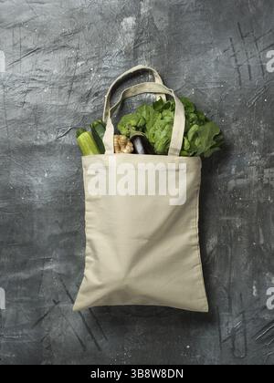 Cotton tote bag with vegetables on gray background. Organic cotton canvas bag with fresh zucchini, eggplant, ginger root, broccoli and salad. Vertical Stock Photo