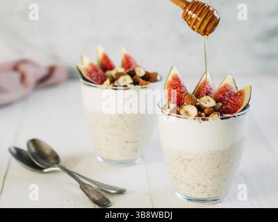 Overnight oats with yogurt, figs and nuts in glass on white wooden table. Ideas and recipes for healthy breakfast, snack or dessert. Honey drips into Stock Photo