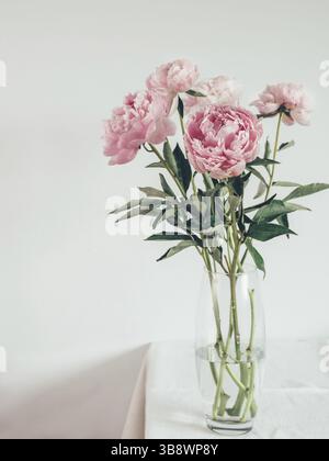 Beautiful pale pink peony bouquet in glass vase on table with white linen tablecloth. Vertical. Copy space for text Stock Photo