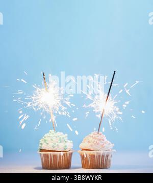 Two cupcakes decorated with colorful sprinkles and sparklers. Festive cupcakes with blue and pink sprinkles on blue background with copy space top for Stock Photo