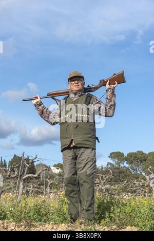 Hunter carrying shotgun on shoulder, wearing hunting vest and cap, standing in a field with grapevines and blue sky in Sant Pere de Ribes, Barcelona, Stock Photo