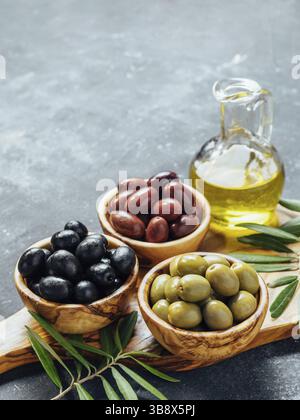 Set of green, black and red or pink olives and olive oil on gray background. Different types of olives in olive wooden bowls and olive oil over wooden Stock Photo